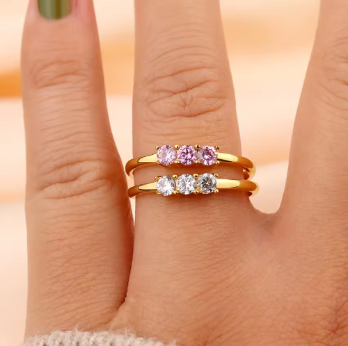 Gold rings with pink and clear gemstones on a finger against a neutral background