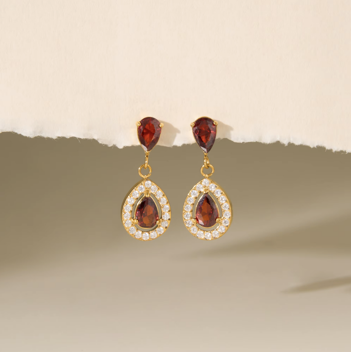 A pair of gold drop earrings with teardrop-shaped dark red gemstones surrounded by small clear stones, displayed against a beige background with a torn paper effect.