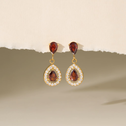 A pair of gold drop earrings with teardrop-shaped dark red gemstones surrounded by small clear stones, displayed against a beige background with a torn paper effect.