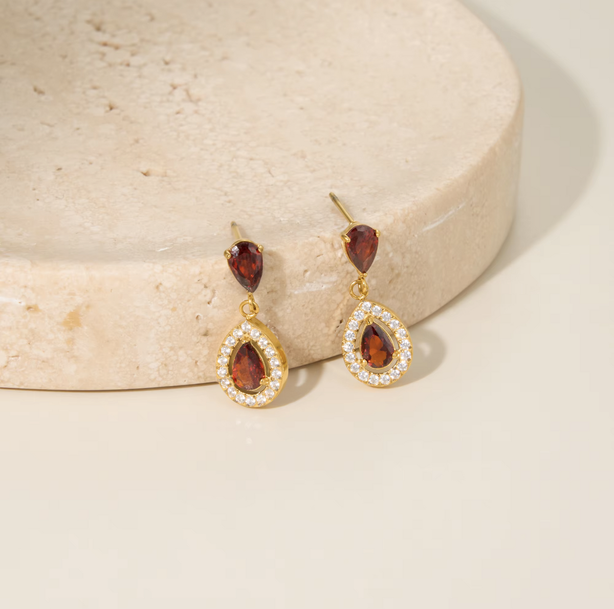 A pair of gold drop earrings with teardrop-shaped brown gemstones surrounded by small clear stones, displayed on a beige stone pedestal against a warm cream background.