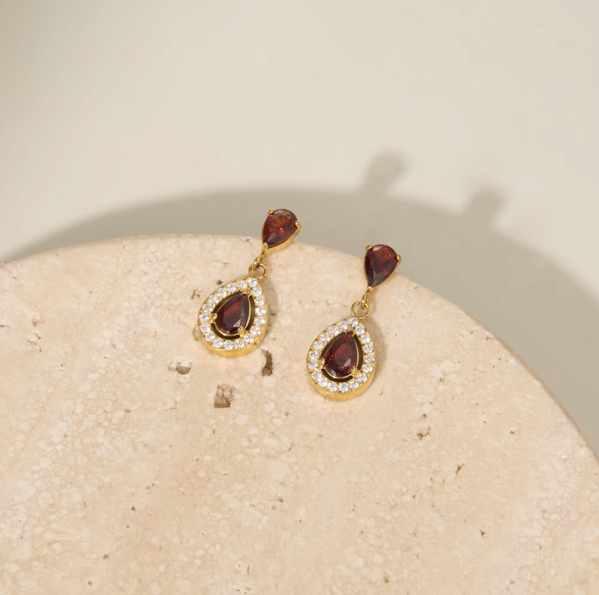 A pair of gold dangle earrings with teardrop-shaped garnet gemstones and a halo of small clear stones, displayed on a textured beige stone surface against a warm cream background.