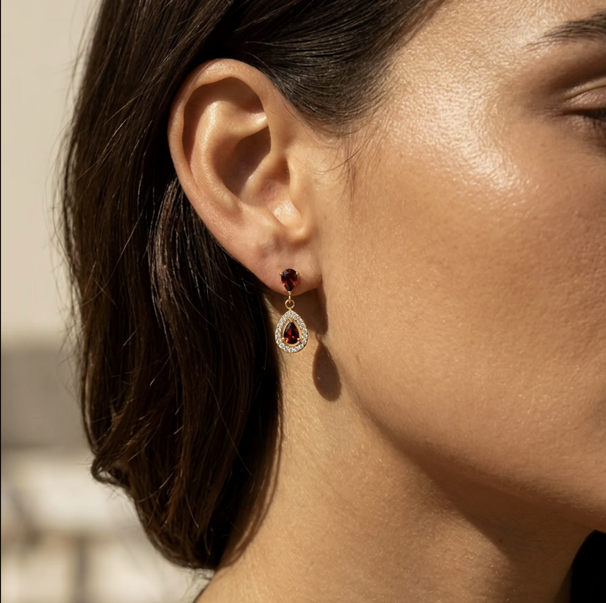 A close-up side profile of a woman wearing a gold drop earring with a small dark red gemstone and a teardrop-shaped red gemstone surrounded by clear stones.