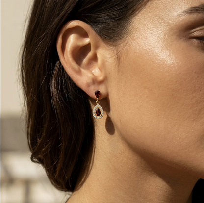 A close-up side profile of a woman wearing a gold drop earring with a small dark red gemstone and a teardrop-shaped red gemstone surrounded by clear stones.
