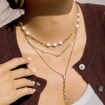 Close-up of a person wearing a gold necklace with pearls and gemstones, with a brown cardigan.