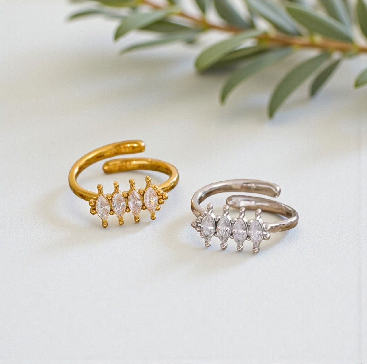 Two adjustable gold and silver rings with gemstones on a light background with greenery.