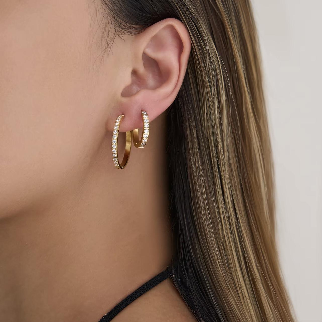 Close-up of a person wearing two different sized gold hoop earrings with diamonds with a neutral background