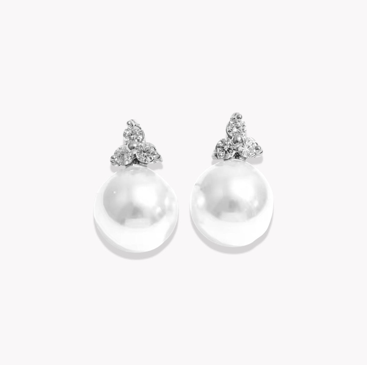 A pair of silver stud earrings featuring a large, lustrous white pearl drop and a small triangular cluster of clear stones at the top, set against a plain white background.