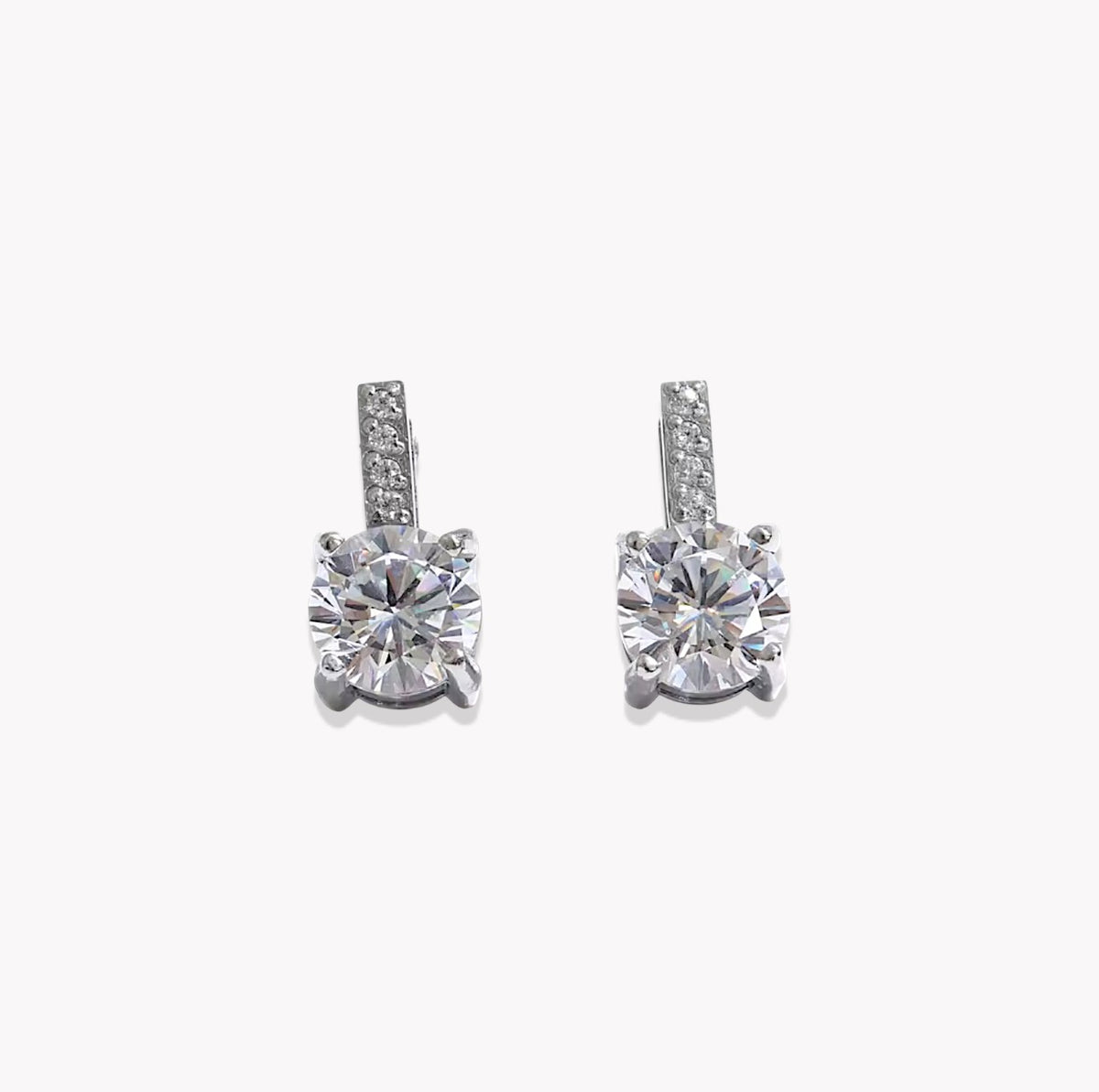 A pair of silver-toned drop earrings featuring a large round-cut clear center stone in a four-prong setting, with a smaller channel-set row of clear stones above it, shown on a plain light gray background.