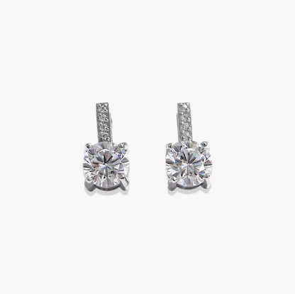 A pair of silver-toned drop earrings featuring a large round-cut clear center stone in a four-prong setting, with a smaller channel-set row of clear stones above it, shown on a plain light gray background.