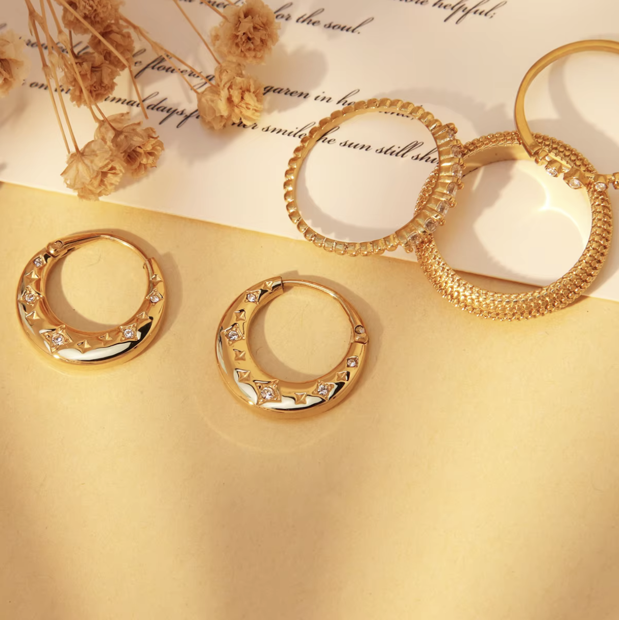 Gold hoop earrings on a textured gold surface with dried flowers and text in the background.