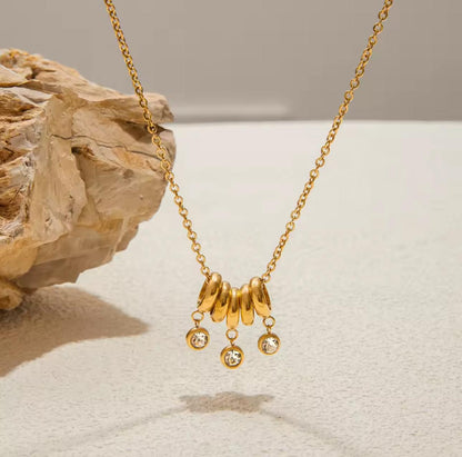gold necklace with 5 rings that move independently of one another. Three of the rings have drop gemstones dangling from them. Pictured on a light grey background