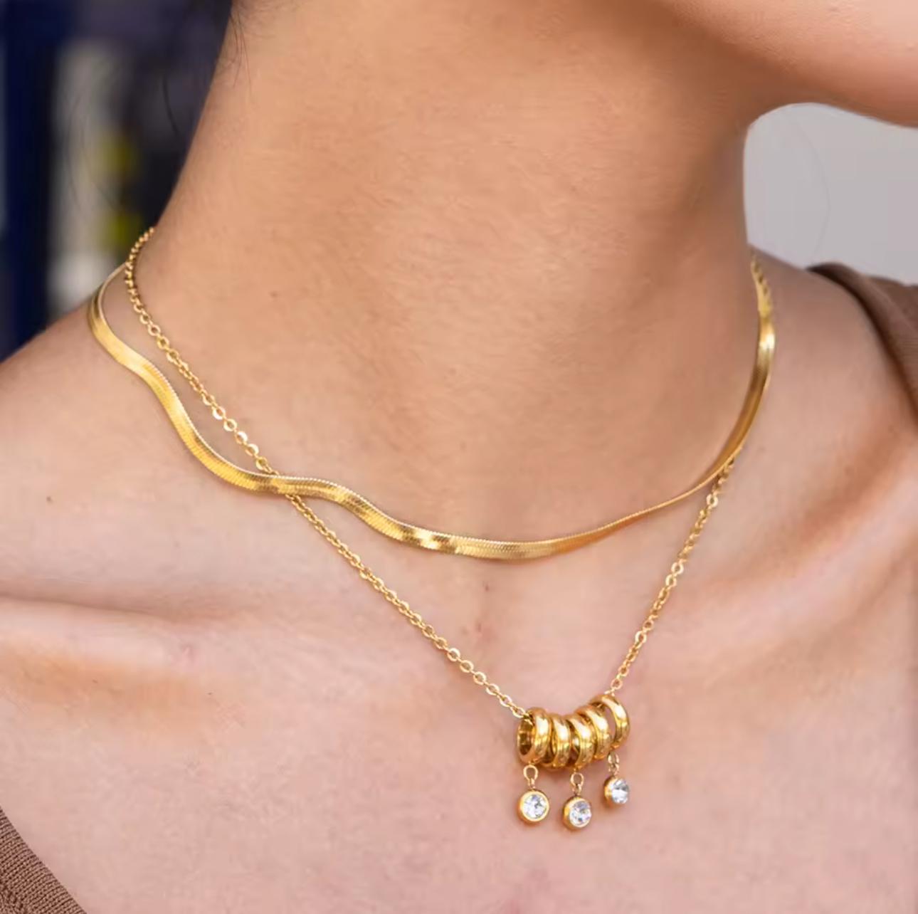 gold necklace with 5 rings that move independently of one another. Three of the rings have drop gemstones dangling from them. Pictured on a neck with another necklace