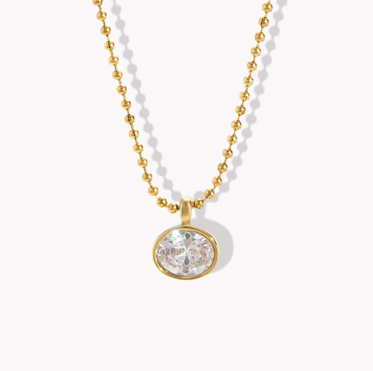 A gold ball chain necklace with an oval bezel-set clear gemstone pendant, shown on a clean white background with soft drop shadows.