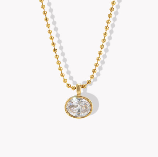 A gold ball chain necklace with an oval bezel-set clear gemstone pendant, shown on a clean white background with soft drop shadows.