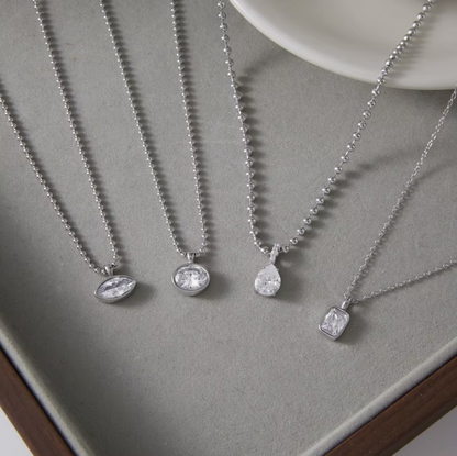 Four silver pendant necklaces with clear gemstones in different shapes, arranged on a gray velvet jewelry tray.