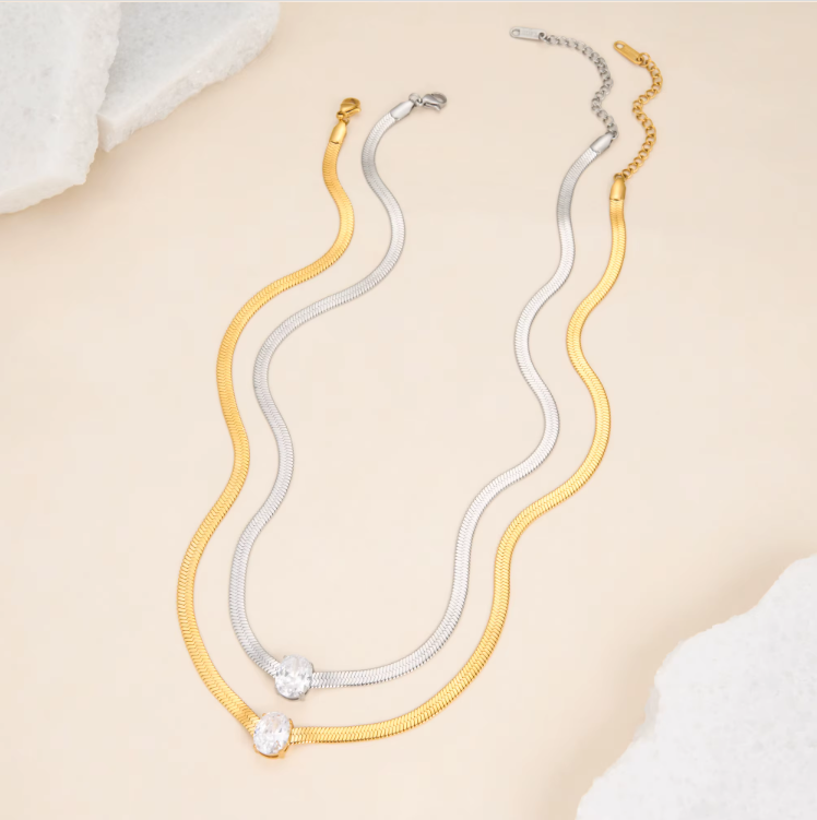 one gold and one silver necklace, both with an oval shaped gemstone pictured together on a light beige background