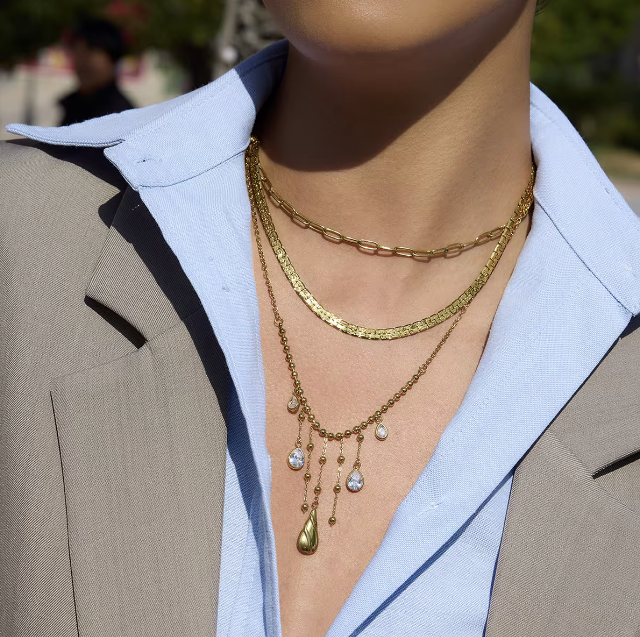 3 gold necklaces layered together. one as a small paperclip necklace, the second is a snake chain and the third is a longer chaing with dangling gemstones. Pictured being worn around the neck