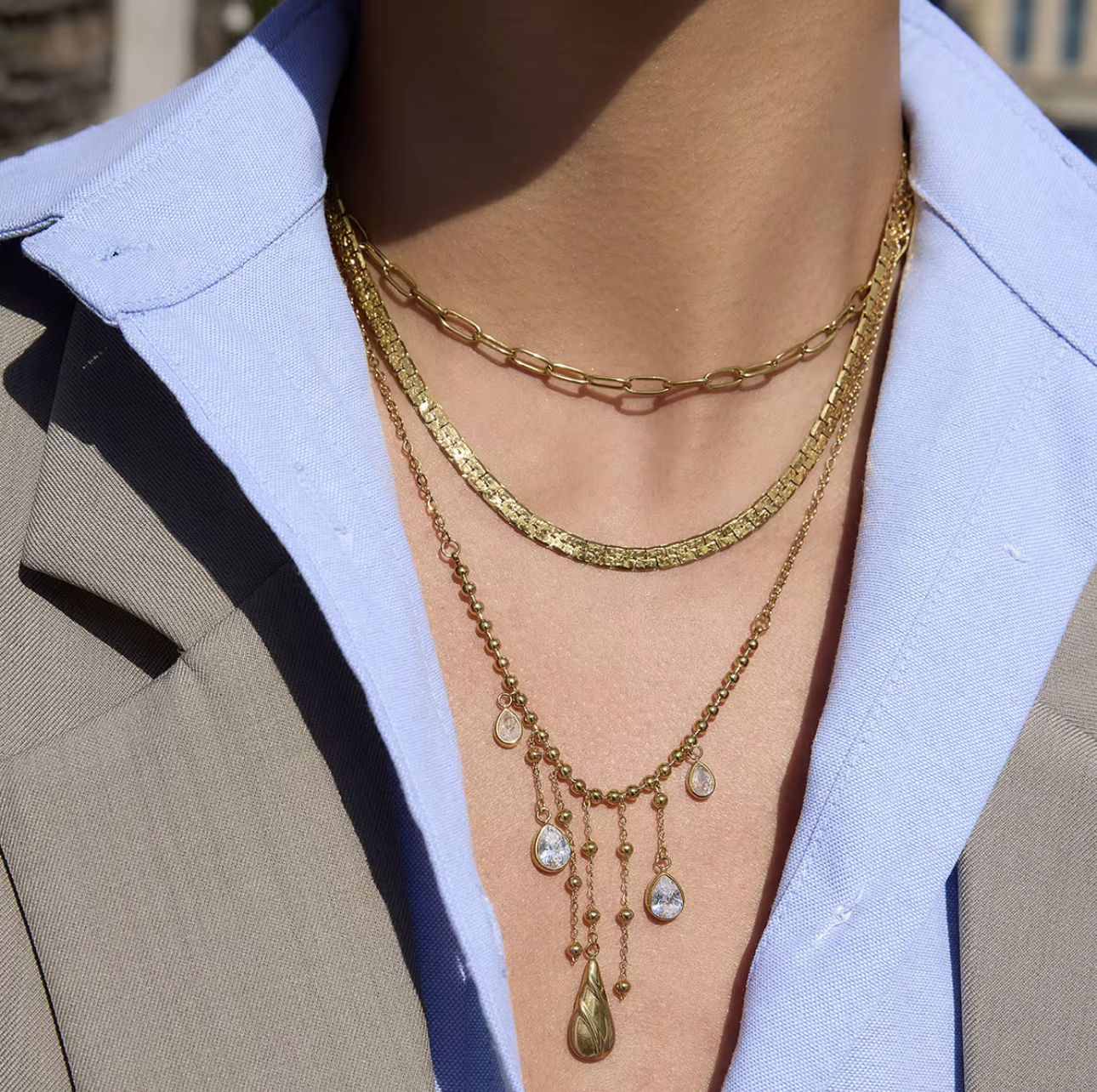 3 gold necklaces layered together. one as a small paperclip necklace, the second is a snake chain and the third is a longer chaing with dangling gemstones. Pictured being worn around the neck