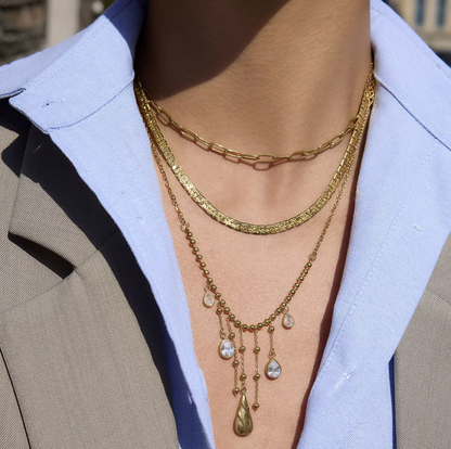 3 gold necklaces layered together. one as a small paperclip necklace, the second is a snake chain and the third is a longer chaing with dangling gemstones. Pictured being worn around the neck