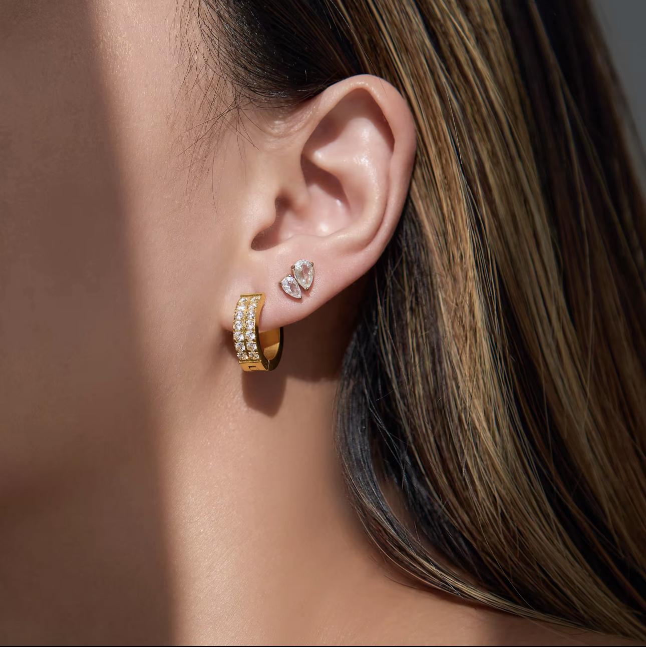 Close-up of an ear wearing gold hoop earrings with clear gemstones.