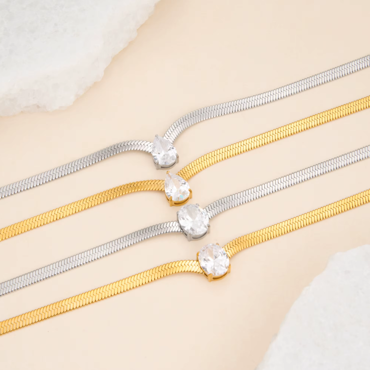 2 silver and 2 gold necklaces with oval and pear gemstones pictured on a light beige background