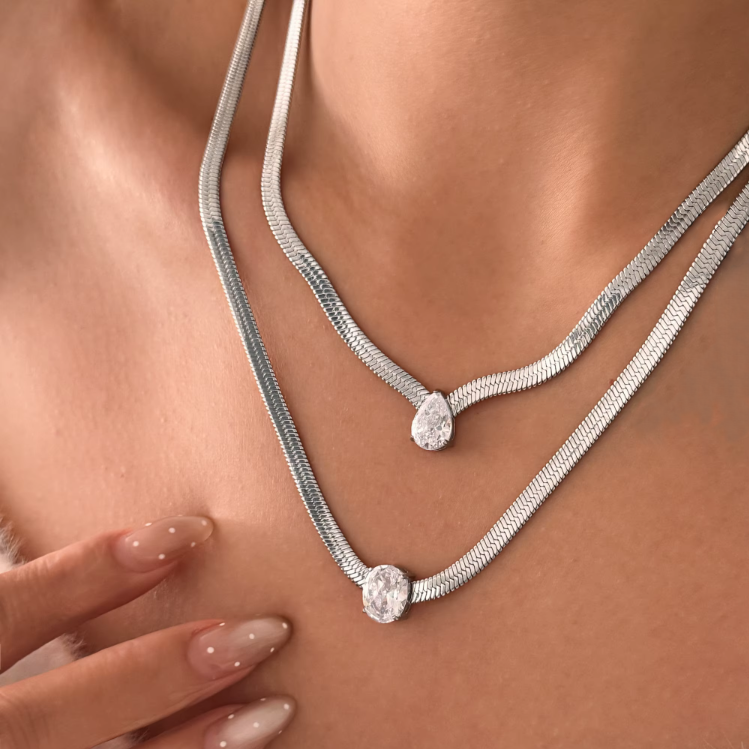 2 silver necklaces, one with a pear pendant and the other with an oval pendant, layered on a persons neck